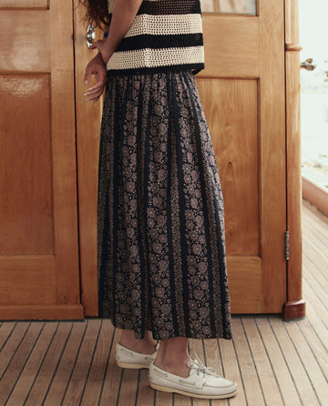 Wearing THE GREAT. SU26’s The Canary Skirt in Black And Cream Token Floral, a person stands indoors; only the lower half of their body is visible.