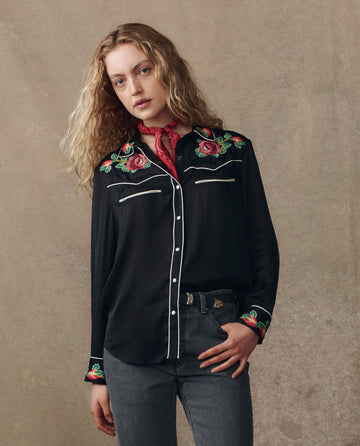 A woman with wavy blonde hair wears THE GREAT. H25 COLLECTION's The Heirloom Rose Western Shirt in black, paired with a rose red scarf and gray jeans, standing against a neutral background.