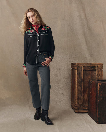 Indoors, a person wears THE GREAT. H25 COLLECTION's "The Heirloom Rose Western Shirt" in black, paired with gray jeans, black boots, and a red bandana. Wooden crates and an heirloom rose complete the background scene.