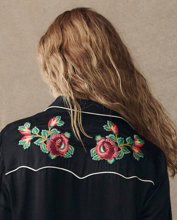 A person with wavy, light brown hair wears THE GREAT. H25 COLLECTION's The Heirloom Rose Western Shirt in black, featuring white piping and red heirloom rose embroidery on the back.