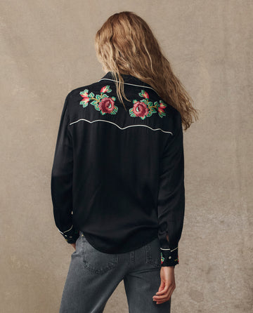 A person with long wavy hair faces away, wearing THE GREAT. H25 COLLECTION's The Heirloom Rose Western Shirt in black with rose embroidery on the back and grey jeans, set against a neutral background.