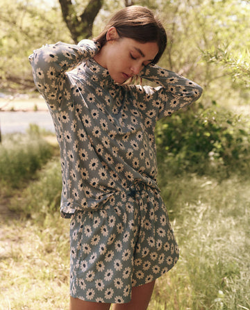 In a grassy outdoor area, a person wears a matching floral top and The Rover Short in Falling Daisy by THE GREAT. SP25 TGO, casually placing their hands behind their neck while looking downward.