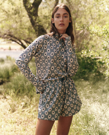 Wearing a vibrant floral outfit, a person enjoys the outdoors, blending seamlessly with nature. Their "The Quarter Zip Trail Pullover - Falling Daisy" by THE GREAT. SP25 TGO enhances the scenery as they stand gracefully among trees and grass.