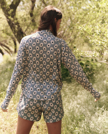 A person in casual wear, featuring a long-sleeve floral shirt and THE GREAT. SP25 TGO's "The Rover Short" in Falling Daisy, stands amidst lush green foliage and towering trees.