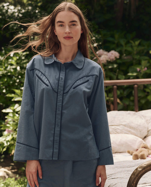 A woman with long brown hair stands outdoors wearing THE GREAT. SP25 SLEEP's The Western Pajama Shirt in Blue Dusk. A bed with pillows and greenery can be seen in the background.