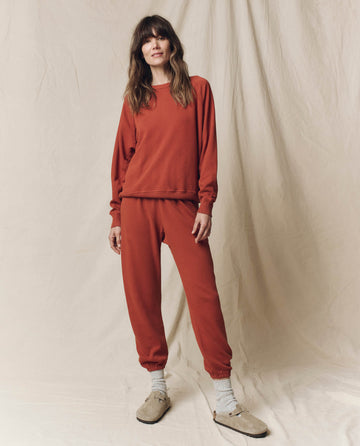 In Los Angeles, a woman stands before a beige fabric backdrop, wearing THE GREAT. F25 SEASONAL KNITS The Stadium Sweatpant. Solid -- Brick, paired with white socks and beige slip-on shoes.