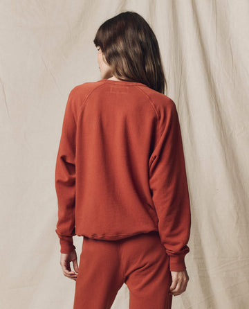 A person with long brown hair, seen from behind, wears THE GREAT. F25 SEASONAL KNITS The College Sweatshirt. Solid -- Brick and matching pants, evoking vintage style, standing against a neutral fabric backdrop.