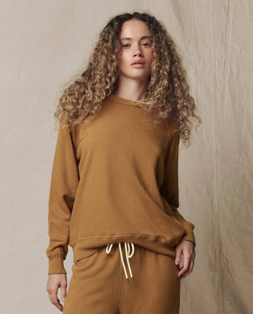 A person with curly hair is wearing THE GREAT. SU26 SEASONAL CORE The College Sweatshirt in Solid -- Cork, standing against a neutral fabric backdrop.