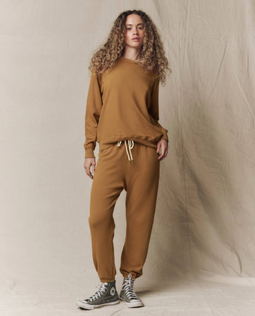 A person with curly hair wears THE GREAT. SU26 SEASONAL CORE The Stadium Sweatpant in Solid Cork, paired with a brown long-sleeve sweatshirt and gray high-top sneakers, standing on a neutral backdrop.