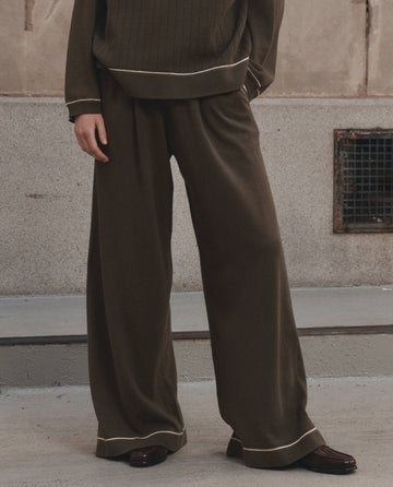A person standing outdoors on concrete steps, with only the lower half of their body visible, wears The Menswear Tipping Trouser Pant in Olive by THE GREAT. F26 D1—loose-fitting menswear pants featuring a dark olive tone and light piping detail.