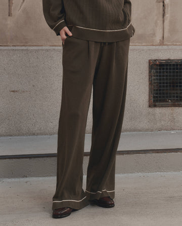 A person wearing THE GREAT. F26 D1 The Menswear Tipping Trouser Pant in olive, paired with a matching sweater featuring white trim, stands on a concrete surface near a wall with a vent.
