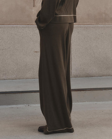 A person from the waist down wears THE GREAT. F26 D1's The Menswear Tipping Trouser Pant in Olive, styled with a matching knit jacket featuring white trim and standing on concrete—an understated take on modern menswear.