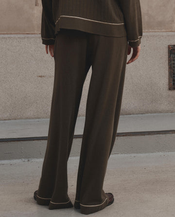 A person stands with their back to the camera, wearing THE GREAT. F26 D1 The Menswear Tipping Trouser Pant in olive, paired with a matching top, on a concrete surface near a beige wall.