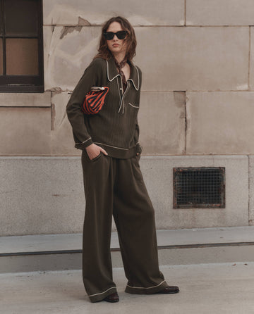 A woman stands against a stone building wearing dark sunglasses, an olive green collared top, a neck scarf, and THE GREAT. F26 D1 The Menswear Tipping Trouser Pant in Olive, with a striped pouch tucked under her arm.