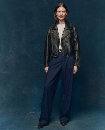 A woman stands against a dark blue background wearing a black leather jacket, white blouse, and THE GREAT. F25 COLLECTORS The Menswear Trouser in Dark Blue Stripe with black shoes.
