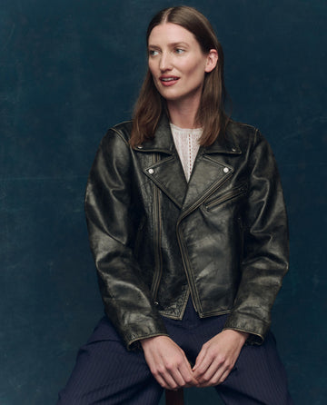 A woman with long brown hair sits on a stool against a dark blue background, wearing THE GREAT. F25 COLLECTORS The Perfect Leather Jacket in Worn Black over a white blouse and dark pants.