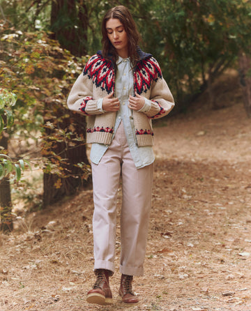 A person wearing THE GREAT. F25 TGO Nordic Sweater—Glacier Fair Isle—along with light pants and boots stands on a forest path covered with fallen leaves.