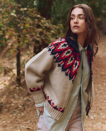 Someone stands outdoors on a dirt path in THE GREAT. F25 TGO's The Nordic Sweater – Glacier Fair Isle, featuring a red and navy Nordic pattern over a light shirt, surrounded by trees.