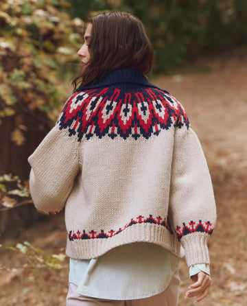 A person with long brown hair walks outdoors wearing THE GREAT. F25 TGO's The Nordic Sweater—Glacier Fair Isle, a cream-colored knit with red, navy, and black geometric patterns.