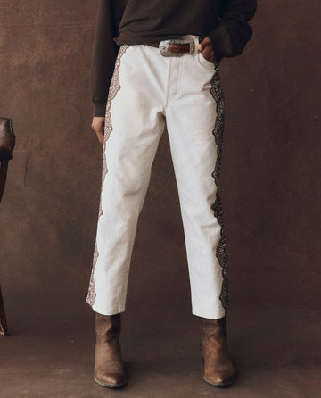 Wearing THE GREAT. F25 D1’s The Lace Print Wayne Jean in cream with brown, paired with a dark brown belt, brown top, and cowboy boots, the person stands against a matching brown background.