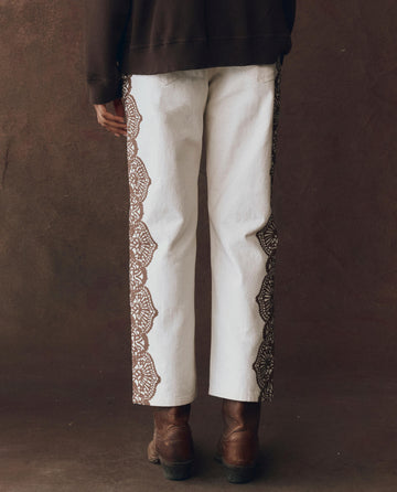 Person wearing THE GREAT. F25 D1 The Lace Print Wayne Jean in cream with brown, featuring a western lace motif on the sides, brown boots, and a dark top, seen from the back against a brown background.