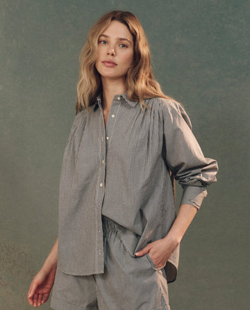 A woman with long hair, wearing THE GREAT. SP25 D2's The Society Top in black and white gingham, poses elegantly against a muted gray background.