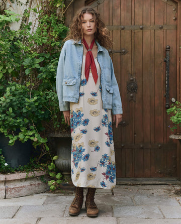 A woman stands before a wooden door, surrounded by green plants, wearing THE GREAT. PF25's The Zip Front Chore Jacket in Dakota Wash over a floral maxi dress, styled with brown boots and a red scarf around her neck.