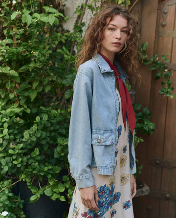A woman with wavy hair stands outdoors wearing THE GREAT. PF25 The Zip Front Chore Jacket in Dakota Wash over a floral dress and red scarf, with green foliage and a wooden door in the background.