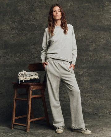 A woman wears THE GREAT. H25 CORE FLEECEBACK The Fleece Victory Sweatpant in Heather Grey, standing by a wooden chair with folded clothes against a textured dark gray background.