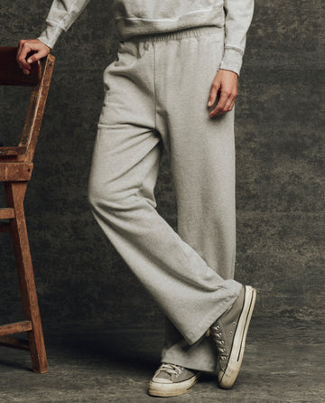 A person wears THE GREAT. H25 CORE FLEECEBACK The Fleece Victory Sweatpant in Heather Grey, standing by a wooden chair against a gray textured background.