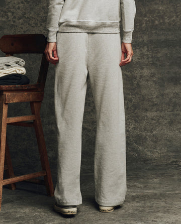 Wearing THE GREAT. H25 CORE FLEECEBACK The Fleece Victory Sweatpant in Heather Grey, a person stands beside a wooden chair with folded clothes, set against a textured gray background.