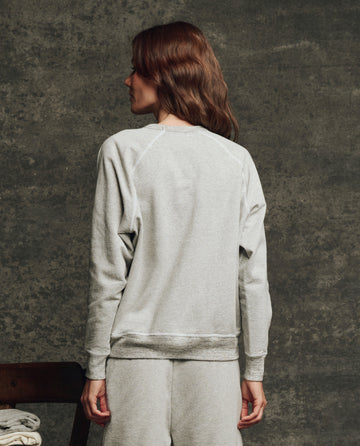 A person faces away, wearing THE GREAT. H25 CORE FLEECEBACK The Fleece College Sweatshirt in Heather Grey, paired with matching sweatpants, set against a textured dark background.