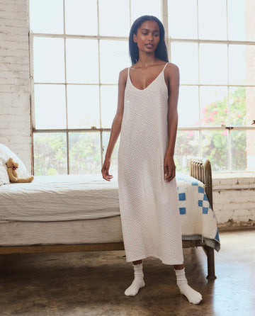 A woman stands indoors on a concrete floor in The Slip Sleeper V Neck Dress in Calico Rose from THE GREAT. PS26 SLEEP, wearing socks beside a bed with striped bedding and a quilt, near large windows.