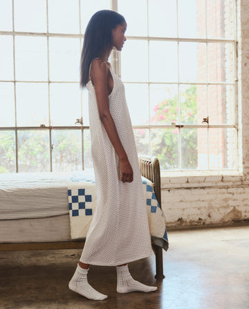 Indoors by a bed, a person wears THE GREAT. PS26 SLEEP’s The Slip Sleeper V Neck Dress in Calico Rose and white socks. Large windows and a quilted blanket appear in the background.