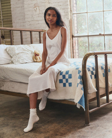 A woman in THE GREAT. PS26 SLEEP The Slip Sleeper V Neck Dress -- Calico Rose, with socks, sits on a bed with a blue and white quilt in a sunlit, rustic bedroom.