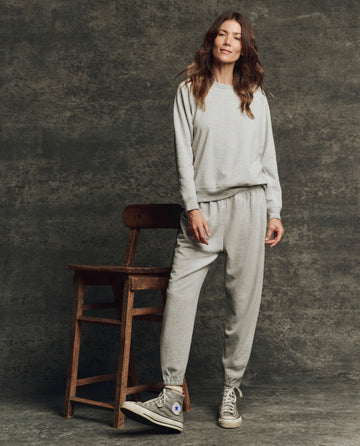 A woman stands by a wooden chair, wearing THE GREAT. H25 CORE FLEECEBACK The Fleece Rookie Sweatshirt in Heather Grey, with matching sweatpants and gray Converse sneakers, posed against a textured gray background.