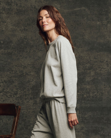 A woman with long brown hair stands sideways wearing THE GREAT. H25 CORE FLEECEBACK The Fleece Rookie Sweatshirt in Heather Grey, paired with matching sweatpants against a textured dark background.