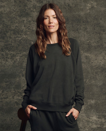 A woman with long brown hair stands against a textured gray background, wearing THE GREAT. H25 CORE FLEECEBACK The Fleece College Sweatshirt in Almost Black, with her hands in her pockets.