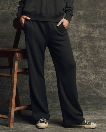 A person stands with hands in pockets wearing THE GREAT. H25 CORE FLEECEBACK The Fleece Victory Sweatpant in Almost Black, paired with a black sweatshirt and gray sneakers beside a wooden chair against a textured gray backdrop.