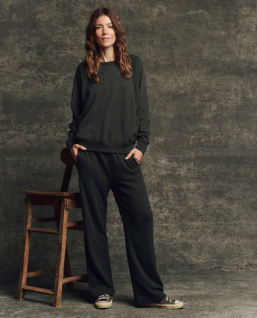 A woman stands beside a wooden chair in THE GREAT. H25 CORE FLEECEBACK The Fleece Victory Sweatpant in Almost Black, paired with a loose black sweatshirt and sneakers, against a textured gray backdrop.
