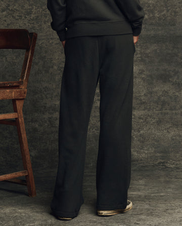 Someone wearing THE GREAT. H25 CORE FLEECEBACK The Fleece Victory Sweatpant in Almost Black and sneakers stands with their back to the camera beside a wooden chair, set against a textured gray backdrop.
