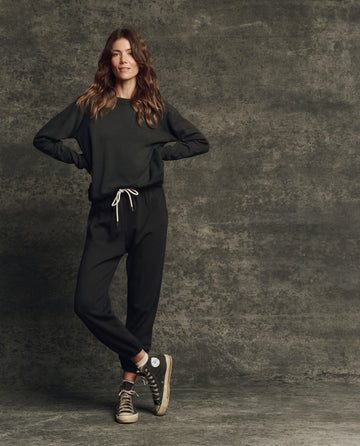 A person stands against a textured gray background wearing THE GREAT. H25 CORE FLEECEBACK The Fleece Stadium Sweatpant in Almost Black, paired with a black sweatshirt and black high-top sneakers.