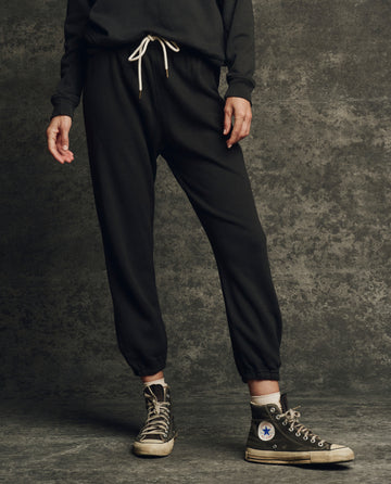 A person stands against a textured background, wearing THE GREAT. H25 CORE FLEECEBACK The Fleece Stadium Sweatpant in Almost Black, a black sweatshirt, and black high-top Converse sneakers.