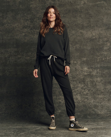 A woman stands before a textured gray background in THE GREAT. H25 CORE FLEECEBACK The Fleece Rookie Sweatshirt in Almost Black, paired with matching joggers and black high-top sneakers.