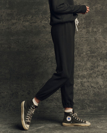 A person wearing THE GREAT. H25 CORE FLEECEBACK The Fleece Stadium Sweatpant in Almost Black, a dark sweatshirt, and black high-top Converse stands sideways against a textured gray background.
