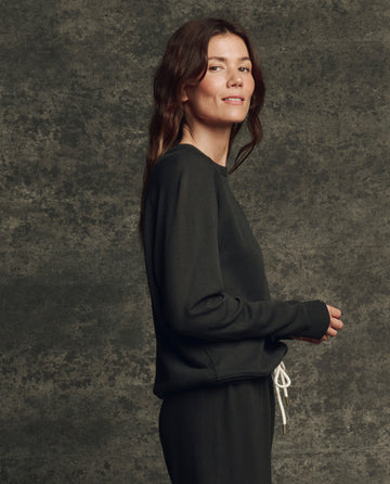 A person with long brown hair stands sideways wearing THE GREAT. H25 CORE FLEECEBACK The Fleece Rookie Sweatshirt in Almost Black and drawstring pants, against a mottled gray background.