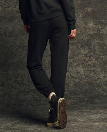 A person stands on one leg against a textured dark background, wearing THE GREAT. H25 CORE FLEECEBACK The Fleece Stadium Sweatpant in Almost Black with a matching sweatshirt and worn black high-top sneakers.