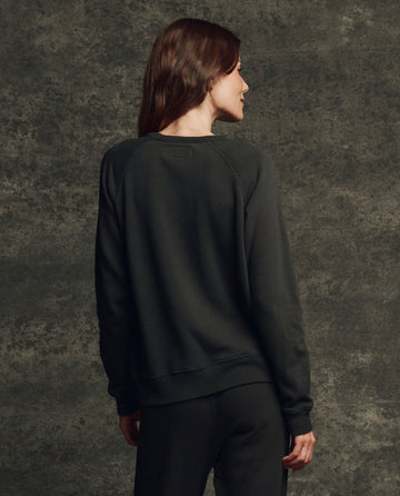 A person stands facing away, wearing THE GREAT. H25 CORE FLEECEBACK The Fleece Rookie Sweatshirt in Almost Black and black pants against a textured dark background.