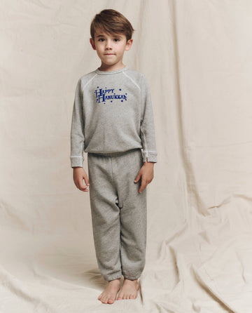 A young child is wearing The Little Stadium Sweatpant in Varsity Grey, by THE GREAT. LITTLE, featuring "Happy Hanukkah" text; they stand barefoot on a beige fabric backdrop.