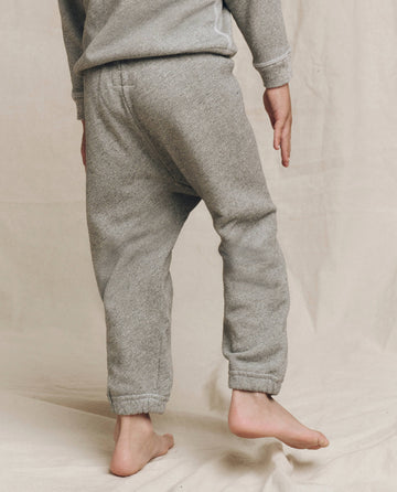 Against a beige background, a child stands on one foot wearing The Little Stadium Sweatpant in Varsity Grey by THE GREAT. LITTLE, paired with a matching gray top. Crafted in L.A., this outfit captures the nostalgic appeal of vintage athletic wear, combining style and playful comfort with ease.